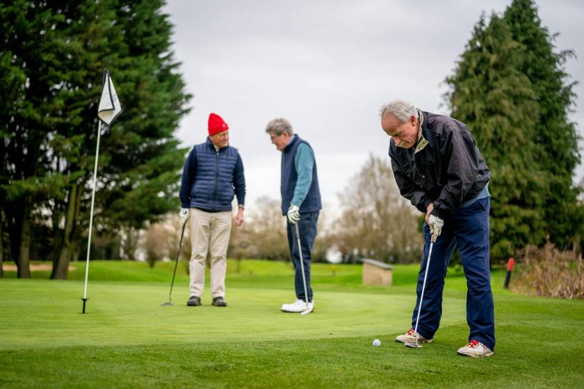 Three Genetlman playing golf. One in foreground putting, two in background talking